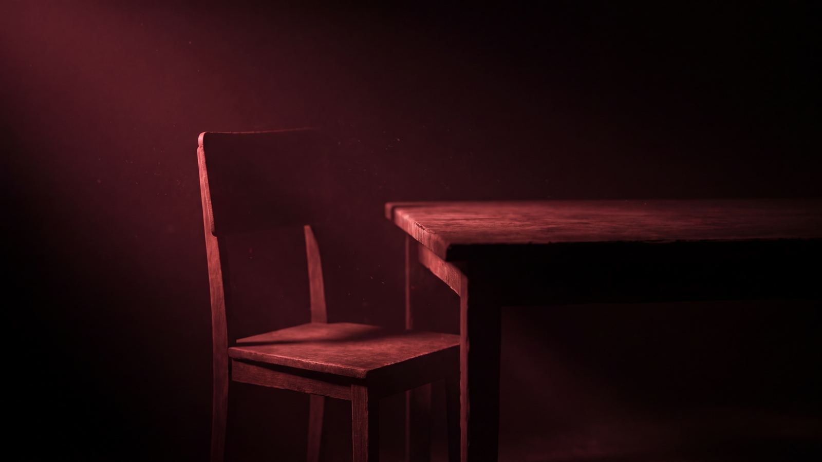 A plain wooden chair beside a bare table in near-darkness, lit from one side in deep crimson.