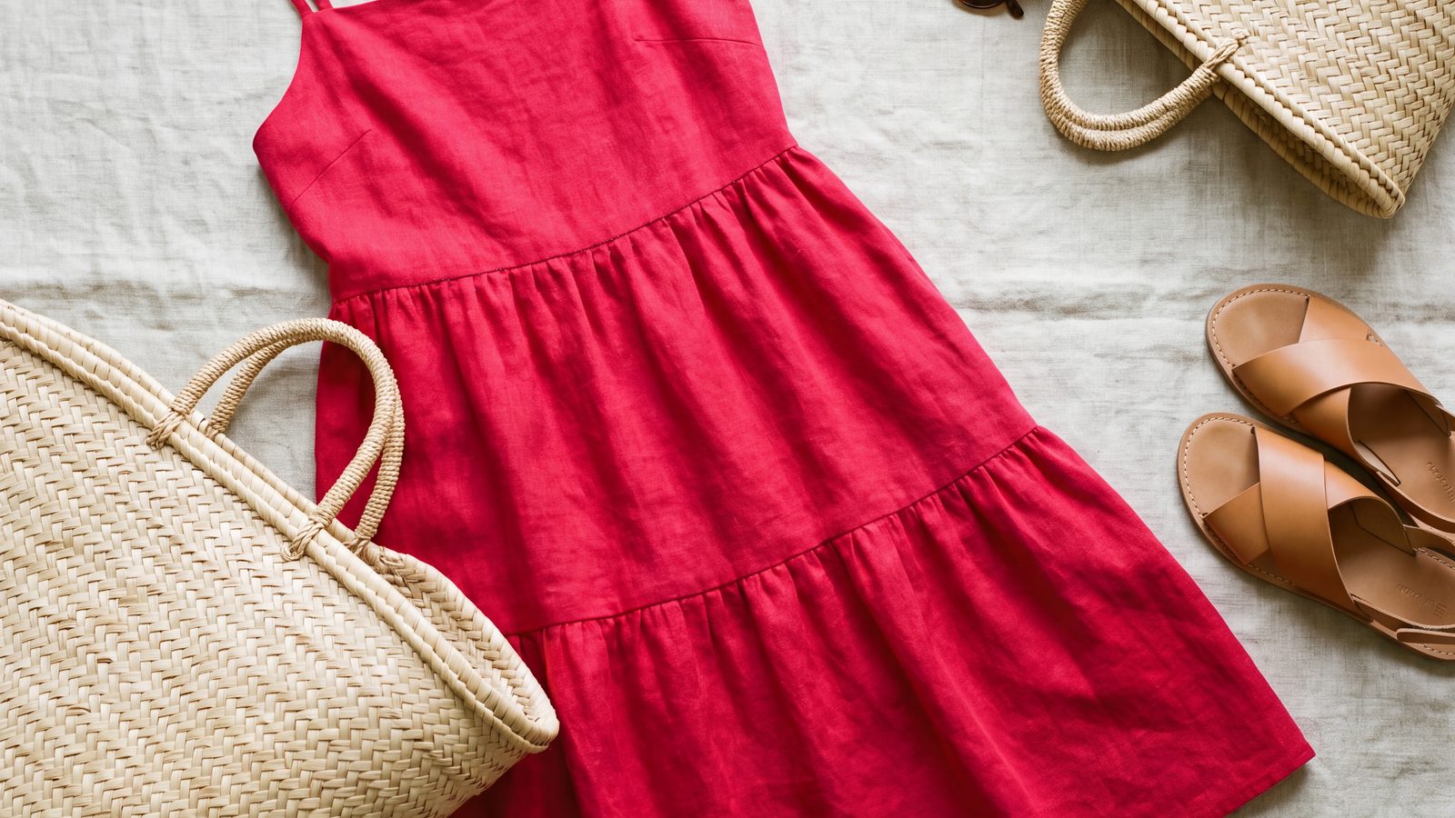 A crimson linen sundress laid flat on oatmeal linen beside a straw tote bag and tan leather sandals.