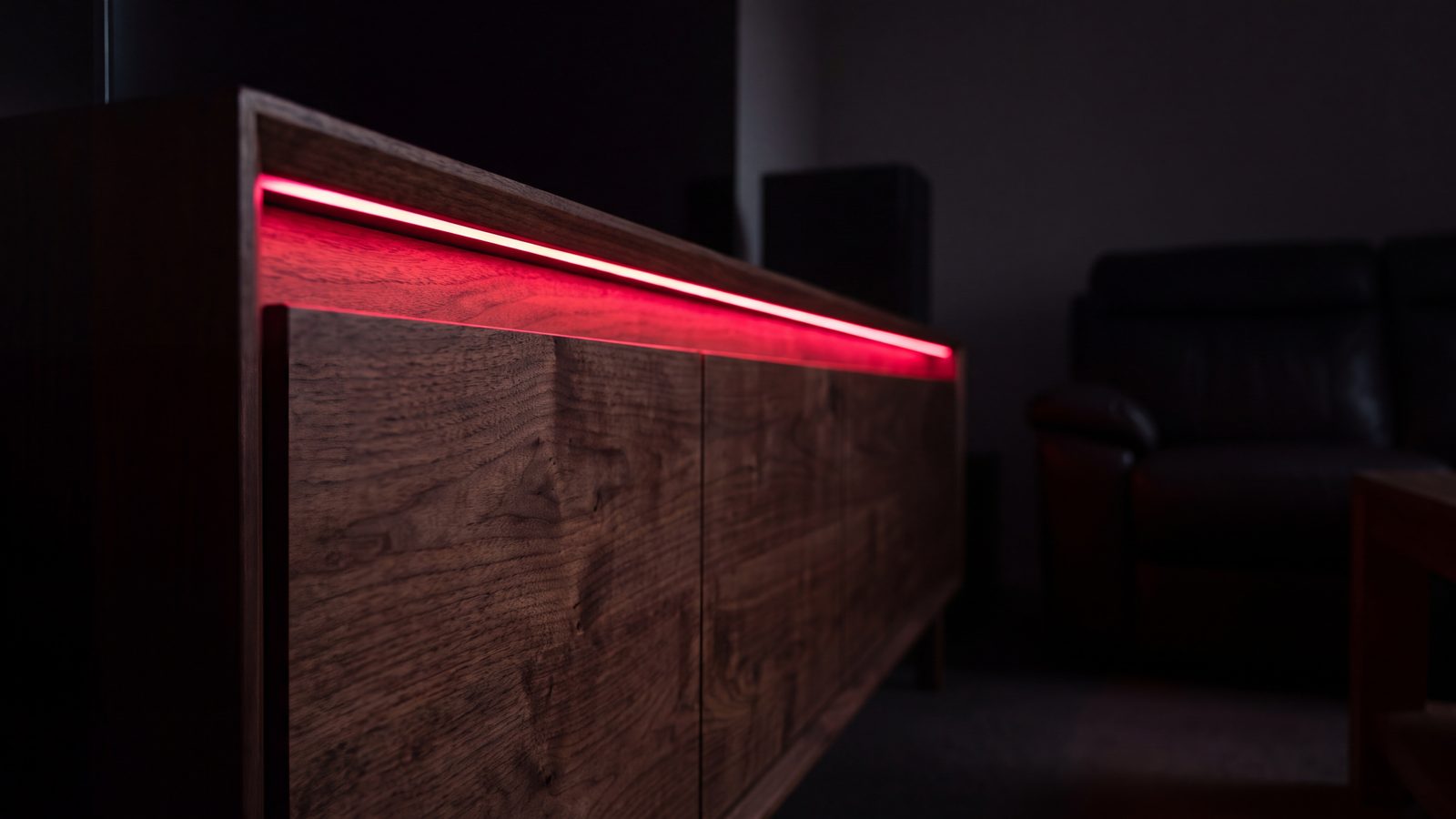 A single run of dark timber cabinetry edge-on in a near-black room, a crimson rim light along its top edge.