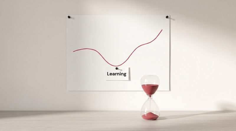 A chart pinned to the wall with a crimson line that dips at a point labelled Learning and then rises, beside an hourglass of crimson sand