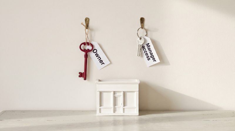 A crimson key tagged Owner and a smaller silver key tagged Manager access hanging on brass hooks above a white model shop front